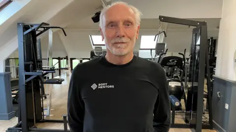 Leeanne Rinne/BBC David Wallis, dressed in gym kit, looks straight into the camera in a weightlifting room.
