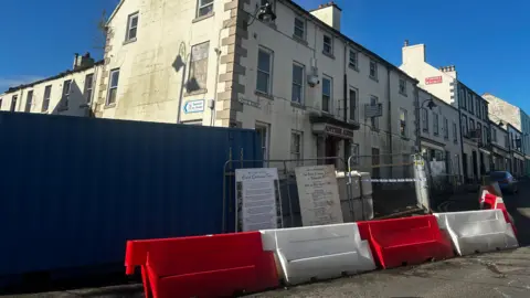 The Antrim Arms Hotel, a white building. Antrim Arms is written in red letters above the door. There are bollards and a fence in front of it.