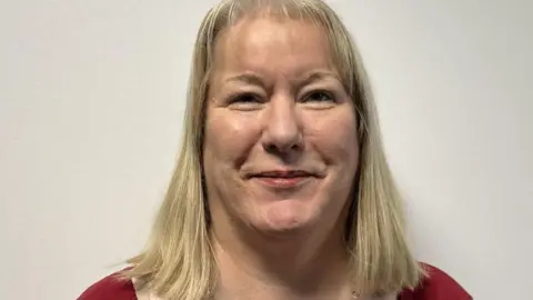 Catherine has shoulder-length blonde hair and is wearing a red top and a long necklace. She is smiling at the camera against a plain white backdrop.