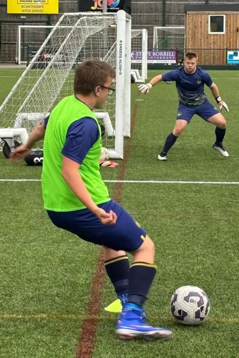 Supplied Taylor in a bright green training bib prepares to kick a ball on an artificial turf pitch while Mason stands ready in front of the goal. Small training goals, fencing, and advertising boards are visible in the background.