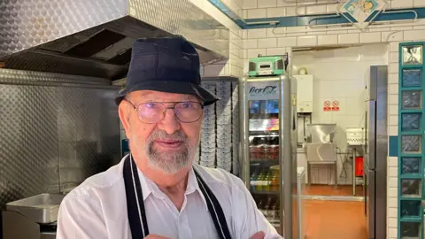 John Paniyas in a white shirt, a black apron and black hat stands in front of frying equipment.