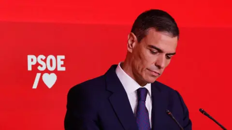 A man in a dark suit and tie looks down with a red backdrop