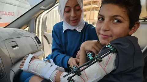 Adam al-Najjar sits in an ambulance with a brace around his arm wearing a dark t-shirt while his cousin, wearing blue and a white headscarf, tends to him.