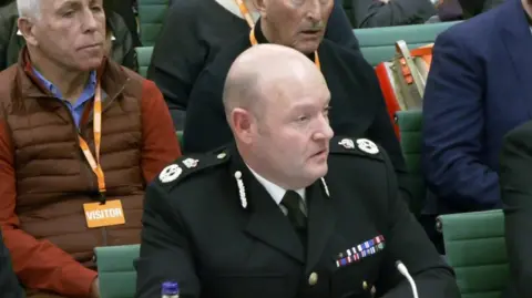 A balding man in a police uniform - black jacket with medals and silver epaulets, wearing a white shirt and black tie sits on a green seat