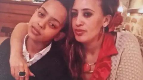 Picture of Biruk on the left, wearing a dark top, leaning into his mum who is wearing a light colour cardigan and red top. She has a red rose in tucked above her ear and has her arm around her son.