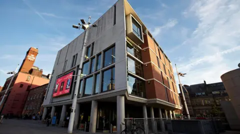 Leeds Museums A modern red-brick building with large glass windows. The building is elevated on cylindrical pillars with an open space underneath. A bright red banner with text and graphics is displayed on the front. In the background are brick buildings and a tower with white lettering which reads 'Electric Press'.
