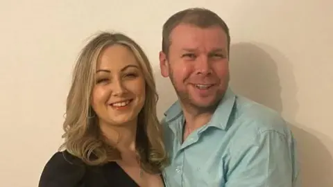 Nicola Byrne Nicola Byrne has long blonde hair and is wearing a black dress. Her partner, Derek, has short brown hair and is wearing a light blue shirt. They are both smiling at the camera.