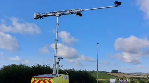 A metal box on the side of the road which is on wheels, it has a large pole rising above it which had cameras looking at an busy country road.