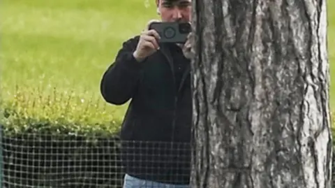 A man in a dark jacket and blue jeans stands partially concealed by a tree trunk and holds a phone up in two hands as if filming and has a white earphone in his right ear 