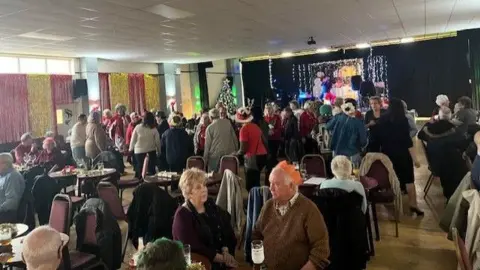 Age UK Hull and East Yorkshire The photo shows a room full of people. Some people are sitting down at tables, with drinks and plates in front of them. Some are standing up, and it looks like they are dancing. Many are wearing party hats from crackers, others are wearing red Santa hats. There is a Christmas tree in the background.