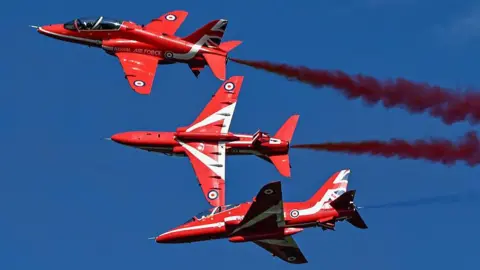 Three Red Arrow jets in mid-air flying next to each. They have red smoke coming out of their engines against a blue sky. The middle one is upside down and performing a rolling manoeuvre.