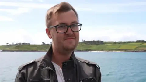 A man with glasses and a leather jacket smiles aboard a ship, with blue sky and blue sea behind him