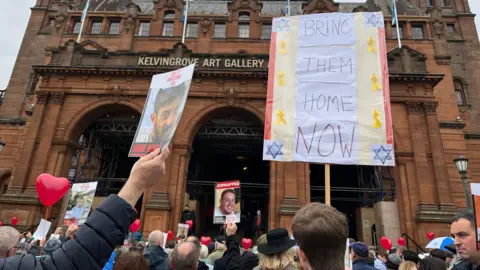 BBC Crowds gathered outside Kelvingrove art gallery. They are holding banners, some show pictures of hostages another has 'bring them home now' written on it.