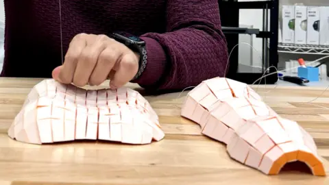 Algorithmic Design Group, MIT-CSAIL Two paper structures sit on a table. They are made of roughly rectangular tiles that are white on the exterior and orange on the interior. 