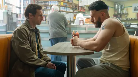 Actors Jamie Bell and Richard Gadd seated opposite each other at a table in a café - it is a scene in Gadd's new series Half Man. Both have serious expressions and are staring intently at each other. Bell wears a khaki jacket and jeans, while Gadd wears a white vest and grey jogging bottoms, a tattoo of a bird on his right shoulder. A man stands at a counter in the background.