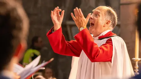 Dale Hodgetts A conductor leading a choir during the opening service of the Three Choirs Festival at Gloucester Cathedral. He is wearing a red top with a white gown and has both of his hands raised in line with his head. His mouth is open, as though he is singing.