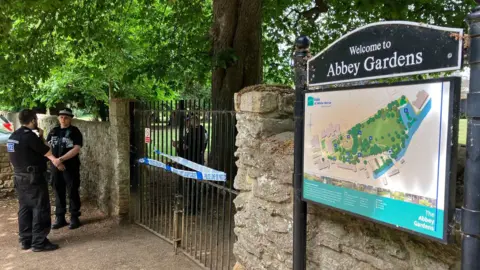 Entrance to Abbey Gardens has blue police tape across it and police officers standing next to it.