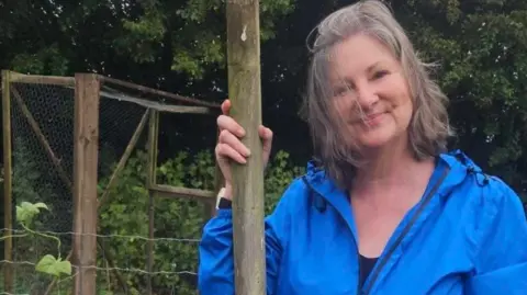 Stuart Smith Pam Wood, a woman with fair hair wearing a blue jacket, standing in front of her allotment plot in Greystoke. In the background there are tall plants growing and she is smiling at the camera.