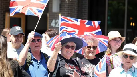 AFP/Getty Images Well wishers wave flags before the arrival of Britain's King Charles III and Queen Camilla at a Lansdowne Park community event in Ottawa, Canada, May 26, 2025.