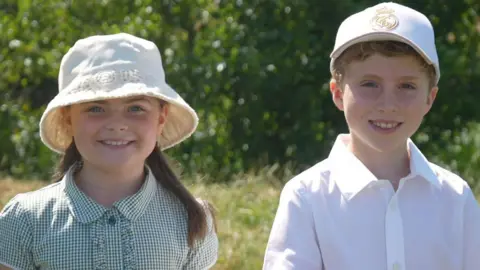 A young girl and boy smile at the camera. They are both wearing school uniform and sun hats and are standing against a backdrop of grass and a riverbed.