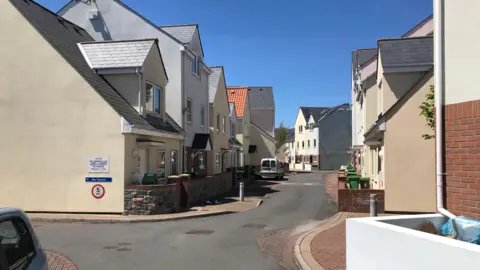 BBC A street in Guernsey with several two-storey and three-storey houses on either side of the road. It is a sunny day and there are no clouds in the sky.