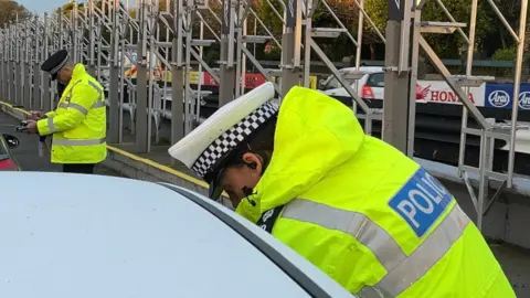 Two police officers in high-vis police jackets lean into cars parked up as park of the checks. One is writing a pad.