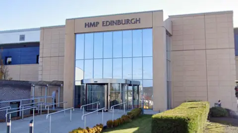 The entrance facade to a prison building, a large glass front and modern beige brick panels above a revolving door. A sign at the top says HMP Edinburgh