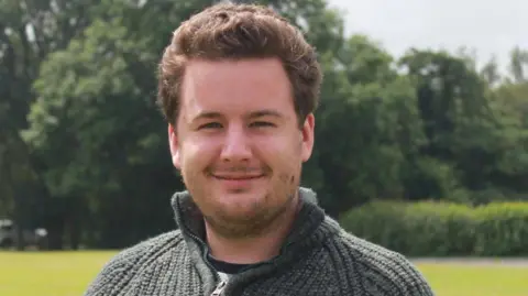 Head-and-shoulders photograph of Thom Campion. He is looking directly into the camera and has dark hair and a thin beard. He is wearing a green jumper and standing in a grassy area with trees in the background.