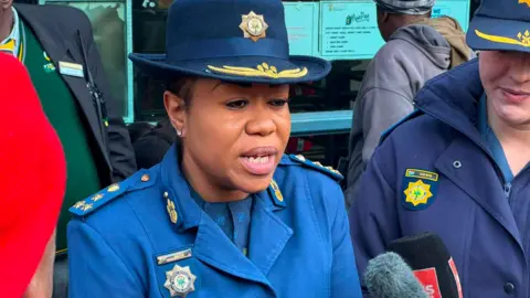 A woman wearing blue police uniform is speaking to gathered media with another officer to her left, and a building behind her.