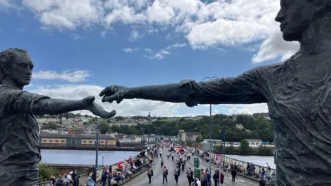 Two statues in the foreground, both of whom are reaching an arm out toward each other. In the background, there is a crowd of people stood on a bridge.