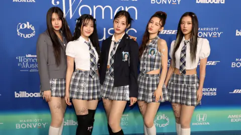 Reuters The five members of NewJeans stand against a blue and turquoise backdrop, dressed in variations of tartan school skirt with blazers and ties. 