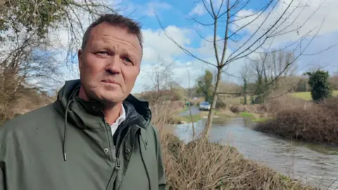 A man in a green coat stands beside a section of the Hampshire chalk stream where vehicles enter and exit