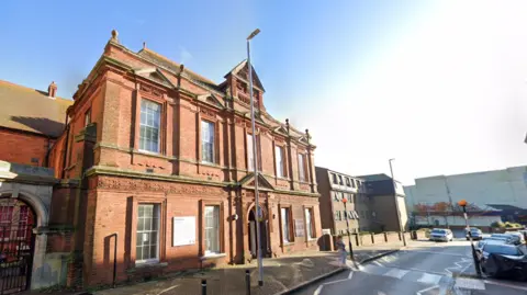 The red brick building on Grace Hill in Folkestone that used to house the town's library. The front of the building is brightly lit and it is a clear day.