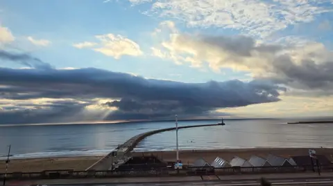 Gill Helps The curved pier at Sunderland which is surrounded by a calm, blue-grey sea. In front of the pier is a beach of dark sand with the pointed roofs of six buildings visible below the road. There is a large grey cloud covering most of the bottom of the sky, but the sky is bright and blue towards the top.