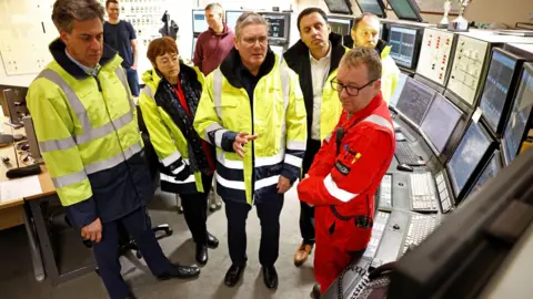 Getty Images Ed Miliband, Sarah Boyack, Sir Keir Starmer, Anas Sarwar and Ian Murray wear an array of hi-vis jackets while staring at a bank of control screens. A man is speaking to them. They are in a control room.