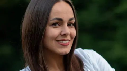 North Yorkshire Council Yana Drapak, from Donetsk, who left Ukraine in 2023. Dana is now an English teacher for North Yorkshire Council - she is smiling towards the camera and has long dark hair and a wide smile and is wearing a white top