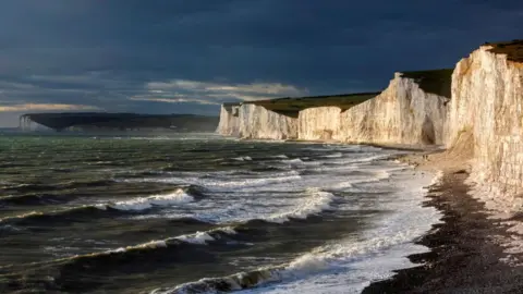 Waves crash to the shore beside some large white cliffs. The sky looks dark and stormy.