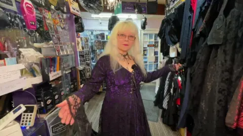 A woman with white hair and glasses, wearing a lace black and purple dress stands in a shop surrounded by goth clothing