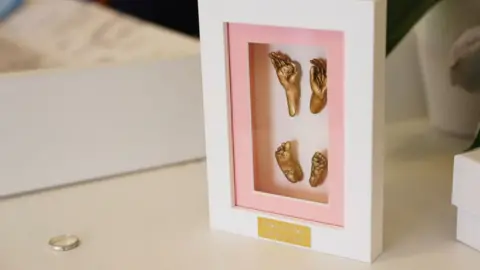 Contributed A white frame that holds golden moulds of a child's hands and feet. A silver ring rests beside it on a white table top.