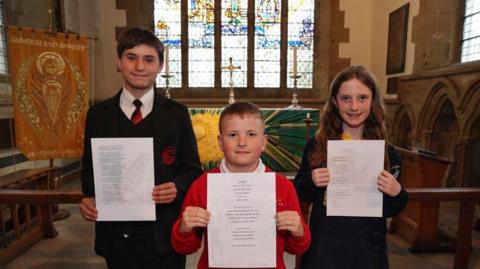 Shaun (left), Lucas (middle) and Milly (right) holding print outs of their poems and smiling at the camera at Sundelrand Minster