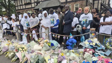 Ibrahima Seck's father clasps his arm around his wife while others stand beside them wearing white shirts with Ibrahima's picture. Floral tributes are laid out on the ground in front of them.