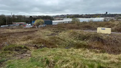 BBC An area of land behind a set of industrial units with a petrol station and Tunstall town centre in the distance.