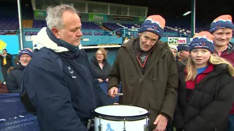 Farmer Richard Messenger holding the drum stick, and is about to bang it on a large white drum. The drum is held by another man, with supporters wearing MND Association hats and holding an MND Association banner around them. Mr Messenger is looking at the camera and smiling. He is wearing a green winter coat and a MND Association woolly hat, which is blue, white and orange. They are inside the Fibrus stadium in Workington.