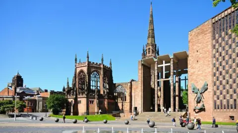 Coventry's old cathedral ruins seen next to the new 20th Century building. There is a clear, blue sky and people are walking around the cathedral precincts. There are decorative globes and a water feature is visible.