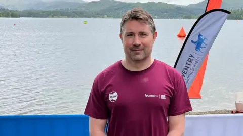 Colin Murphy is standing in front of Windermere with a flag to his right. He is in his thirties wearing a maroon tshirt and has short hair. 