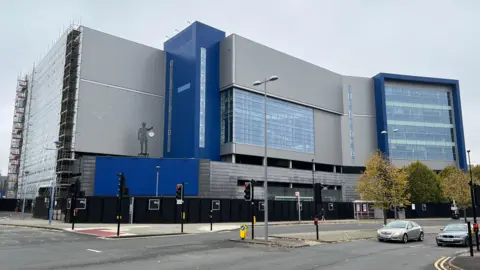 A large grey and blue clad building, which has a large glass section in the centre and another to the right of the building. It sits in front a road junction with traffic lights, some cars are visible on the road