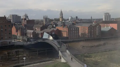 An aerial view of Scale Lane swing bridge, pedestrians can be seen crossing the bridge, with the muddy banks of the River Hull on display. The city's skyline can be seen in the background.