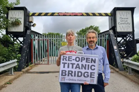 LDRS A man with short hair wearing a blue shirt and blue trousers, and a woman with short hair wearing a grey T-shirt with a red and white logo and blue jeans. She has a bracelet and a necklace. She is holding a sign that reads "Re-open Titanic Bridge, online petition". The bridge is behind them with a tarmac surface, gates, barriers, trees and a sign that read "WP Please Drive Carefully WP".
