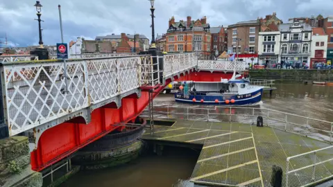 Whitby swing bridge, with white railings and a red base, with a boat passing underneath and shops and other buildings in the background.