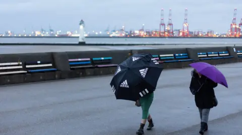 EPA Two women holding umbrellas over their heads shelter fromthe gust and rain on the promenade. A white lighthouse and red tower cranes can be seen in the background on the River Mersey with their lights twinkling in the dark sky.